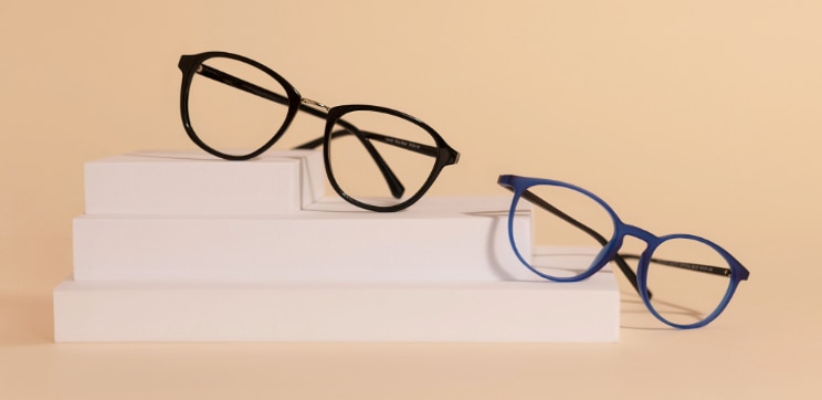 Two pairs of GD Collection round acetate glasses displayed on white geometric blocks — one in shiny black, one in bright blue — against a warm neutral background