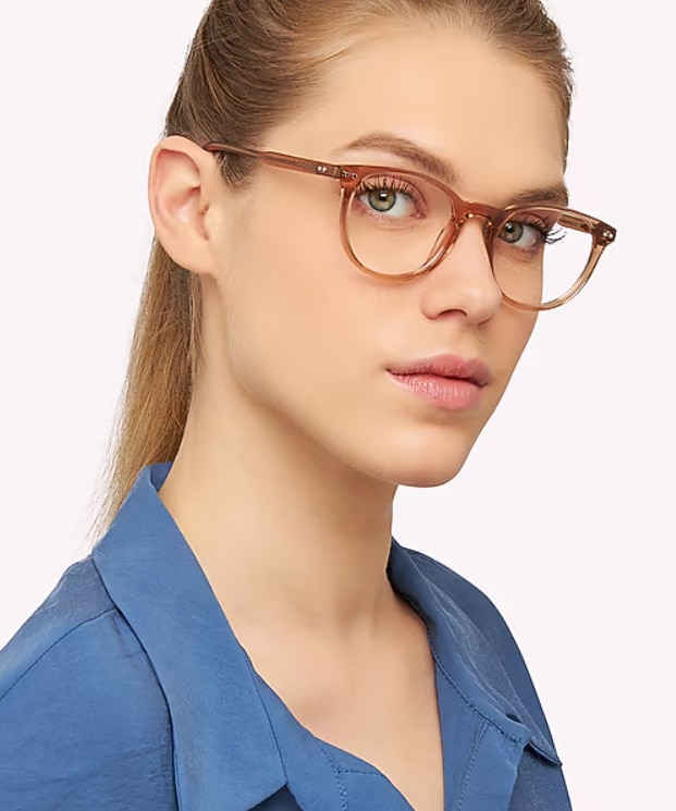A close-up portrait of a woman wearing Scout Dallas translucent light brown phantos-shaped glasses against a light neutral background. The frame has a soft, transparent acetate finish that complements the woman's light features. With her long hair pulled back and dressed in a blue collared blouse, the portrait highlights the eyewear’s modern, refined design and everyday elegance.