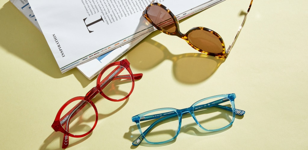 Three pairs of eyewear are arranged on a light-coloured tabletop beside open magazines. Scout Jessica Havana cat-eye sunglasses with brown lenses and metal temples sits at the top, while GD Jessie bright red round glasses and translucent blue rectangular New Balance glasses are positioned below. Strong natural light casts distinct shadows, creating a vibrant, summery lifestyle scene.