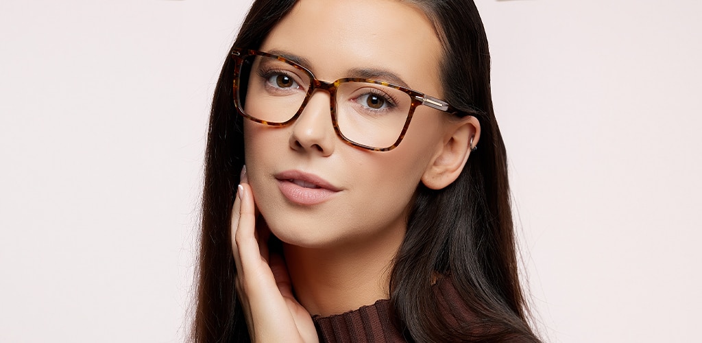 Close-up portrait of a woman wearing London Retro Balham Havana eyeglasses with square frames and gold metal accents along the temples. She has long dark hair and is looking directly at the camera against a light neutral background. The warm brown and amber tones of the frames complement her features.