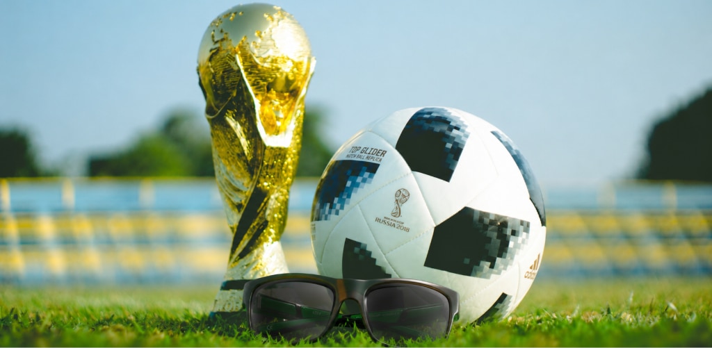 A pair of dark rectangular sunglasses rests on green grass in the foreground of a football pitch. Behind the sunglasses sits a black-and-white football, with a gold World Cup-style trophy standing to its left. The background shows a blurred stadium and trees beneath a clear blue sky. Bright sunlight highlights the trophy’s reflective surface and creates a vibrant, sporty atmosphere.