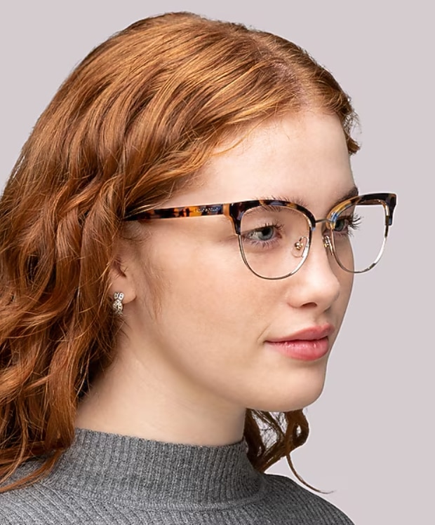 Woman with curly red hair wearing kate spade new york Paityn/G browline glasses with a light tortoiseshell acetate upper frame and gold metal lower rims, against a neutral background.