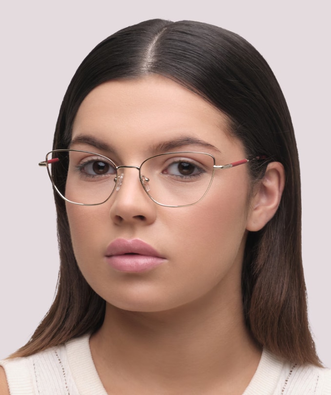 Woman with straight, medium-length dark hair wearing Aspire Heidi gold metal cat-eye glasses with pink accents and a white knitted vest on a plain background.