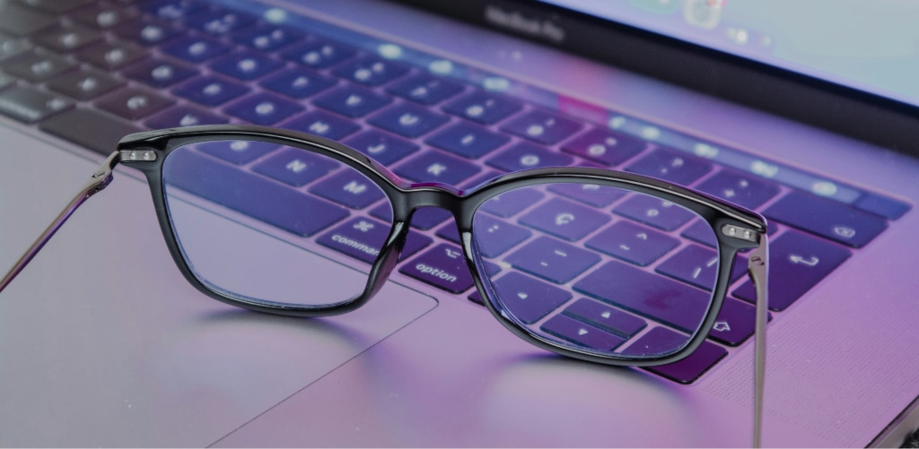 Pair of rectangular black glasses resting on the keyboard of a laptop, with a subtle blue light reflection visible on the lenses and purple-toned ambient lighting across the keyboard.