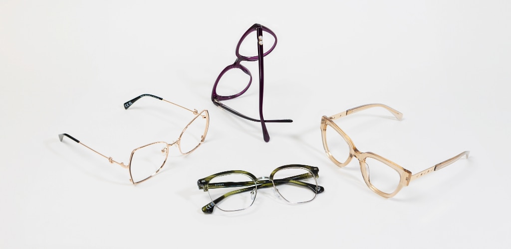 Four pairs of glasses arranged on a white surface, including Scout Kenna gold butterfly metal frames, Scout Kyro purple cat-eye frames, Scout Keegan green square browline frames and Scout Kenzie transparent beige squared cat-eye frames with embellished metal temples, photographed from above with soft shadows.