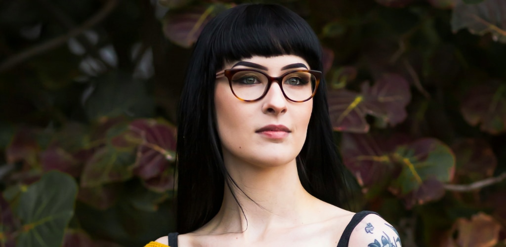 Woman with dark blunt-fringe hair wearing round tortoiseshell glasses, standing against a leafy backdrop — vamp-core eyewear inspiration.