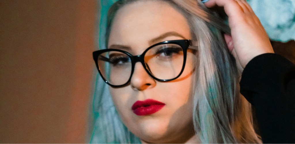 Close-up of a woman wearing bold black cat-eye glasses with red lipstick, hand in her hair, against a moody teal-toned background — gothic-glam eyewear inspiration.