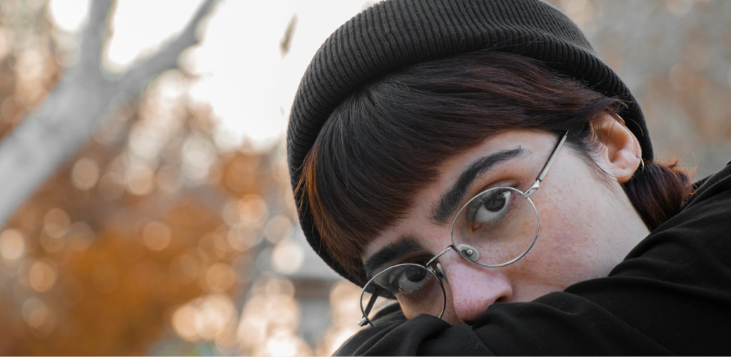 Person wearing a black beanie, round wire-frame glasses and a black top, peeking over their arm against a blurred autumn backdrop — 90s-chic eyewear inspiration.