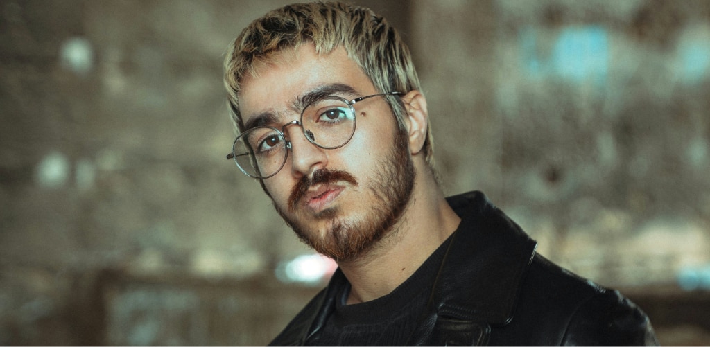Man with dyed blonde hair and a beard wearing round wire-frame glasses and a black leather jacket, against a blurred urban background — grunge-revival eyewear inspiration.