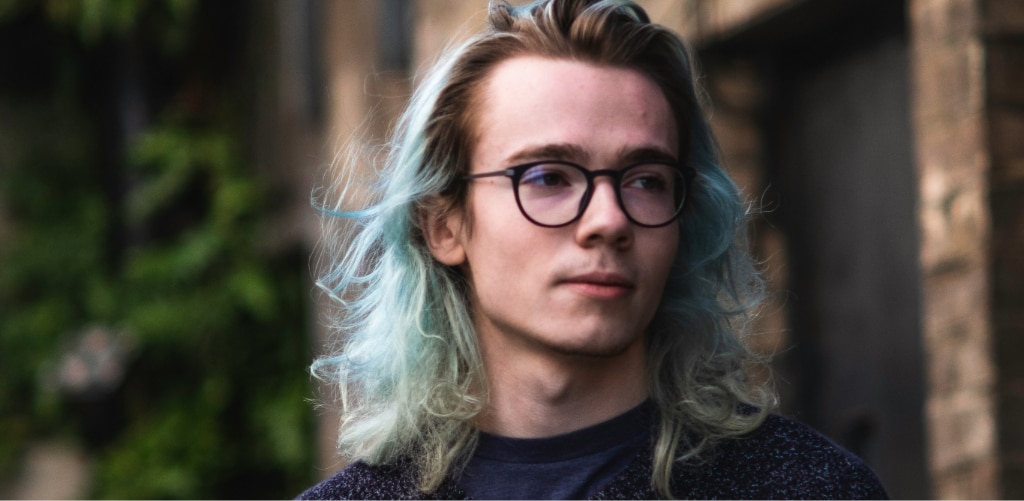 Man with long blue-highlighted blonde hair wearing round black glasses and a dark navy sweater, standing against a blurred urban backdrop — grunge-revival eyewear inspiration.