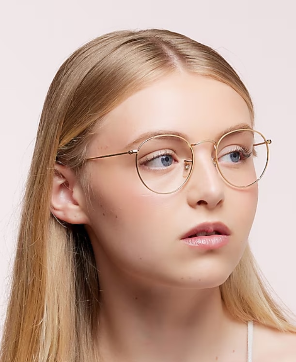 Studio portrait of a young woman with long blonde hair wearing round gold wire-frame Ray-Ban® glasses, looking off to the right against a light grey background.
