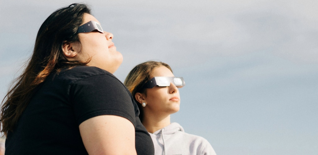 Two people wearing certified eclipse glasses to view the Sun safely.
