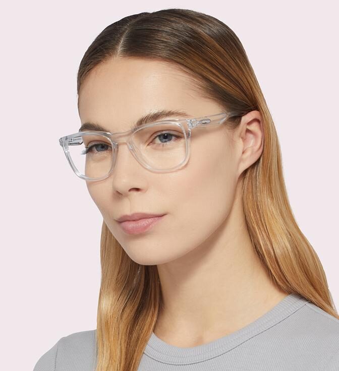 Studio portrait of a woman with long blonde hair wearing clear, rounded square Oakley Trillbe™ X eyeglass frames and a light grey top. She faces slightly toward the camera with a neutral expression against a pale grey background.