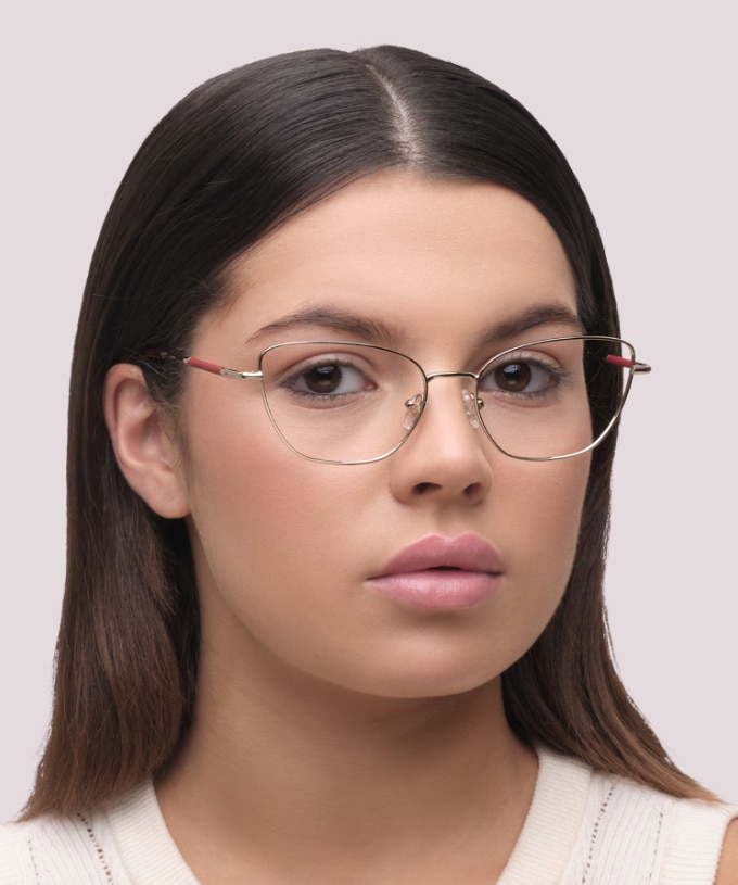 Woman with long, straight, dark brown hair, wearing Aspire Heidi slim gold cat-eye glasses with red temple accents and a cream woollen sleeveless top, looking directly at the camera against a softly blurred light grey background.