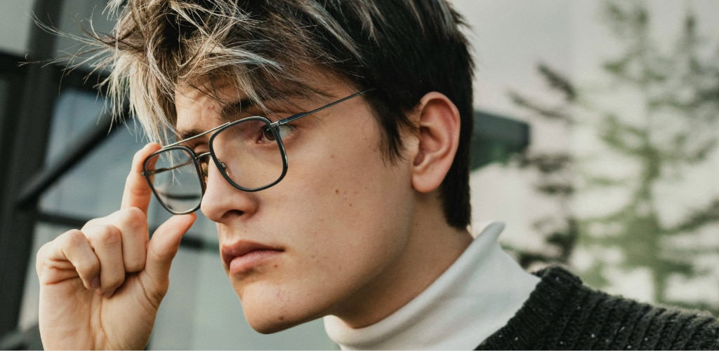 Man with dark hair and blonde highlights wearing black metal-framed square aviator glasses with a white polo neck over a dark knit jumper.