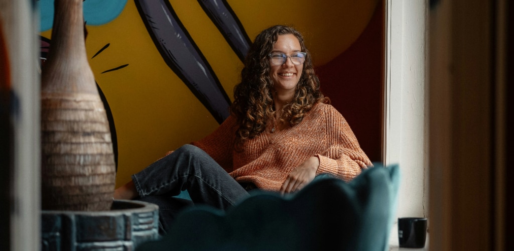 Woman with brown curly hair wearing clear glasses and an orange chunky knit jumper, sitting in a colourful living room.