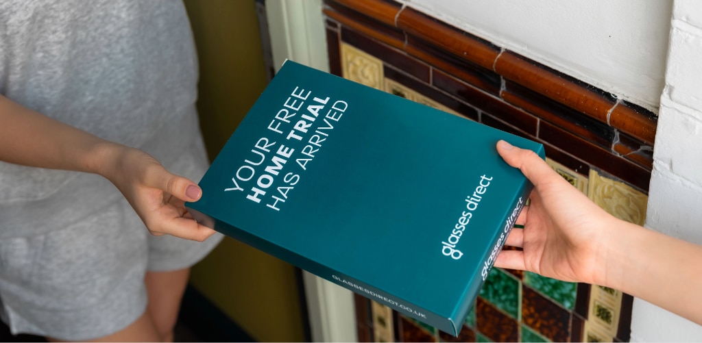 A hand is delivering a teal Glasses Direct box printed with 'Your free home trial has arrived' to someone at their front door.