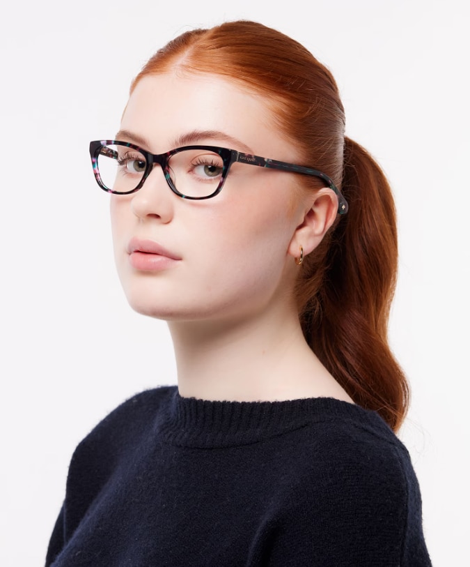 Woman with red hair tied back in a ponytail, modelling Kate Spade Posi rectangular pink-and-green Havana acetate glasses with a navy knitted jumper on a plain background.