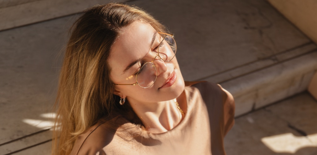 Woman with long blonde hair wearing round gold metal glasses in warm sunlight, with a minimalist neutral outfit.