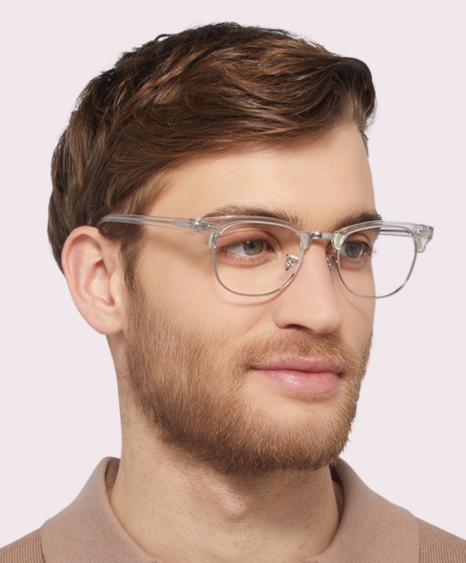 Man with light brown swept hair and a neat beard, wearing a beige knitted polo and Ray-Ban® Clubmaster transparent-and-silver glasses on a plain background.