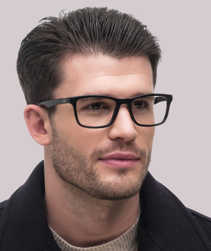 A man with dark hair swept back wearing Ray-Ban RB5279 square black acetate glasses, a beige knitted top and black coat on a plain background.