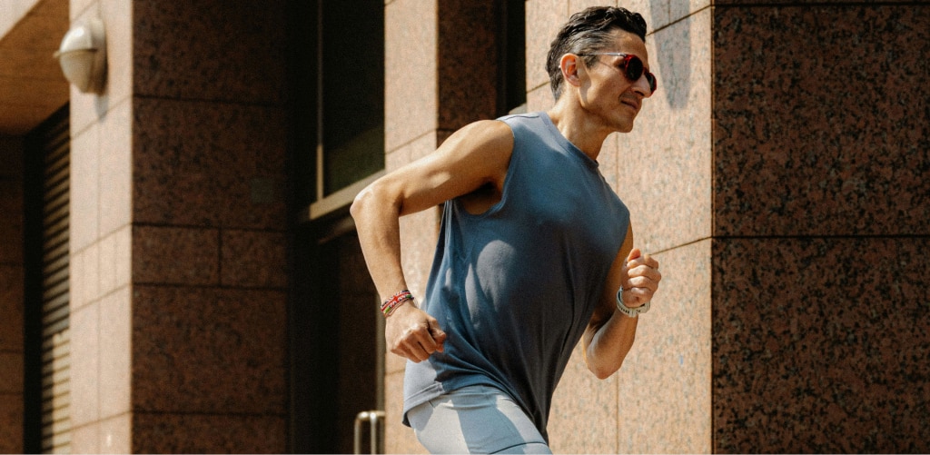 A man with short brown hair in a sleeveless top runs past stone pillars, wearing dark wraparound sunglasses.