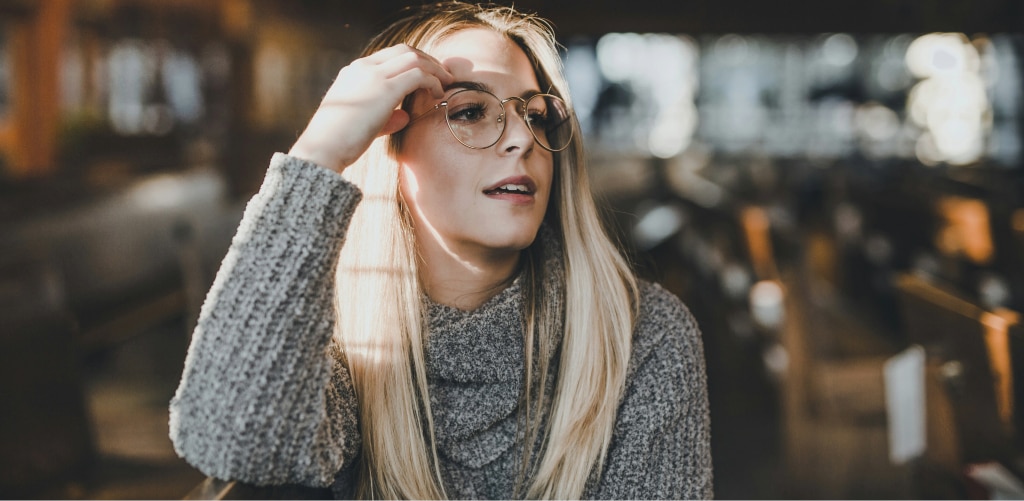 Woman with long blonde hair wearing round metal glasses and a chunky grey knit jumper in an indoor setting.