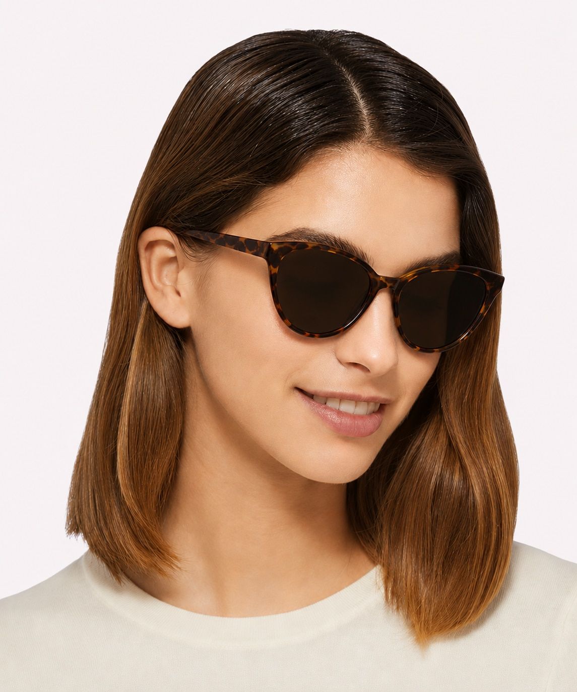 A smiling woman with medium brown hair wears Pink Ribbon Primrose cat-eye Havana sunglasses and a white t-shirt, posed against a clean white studio background.