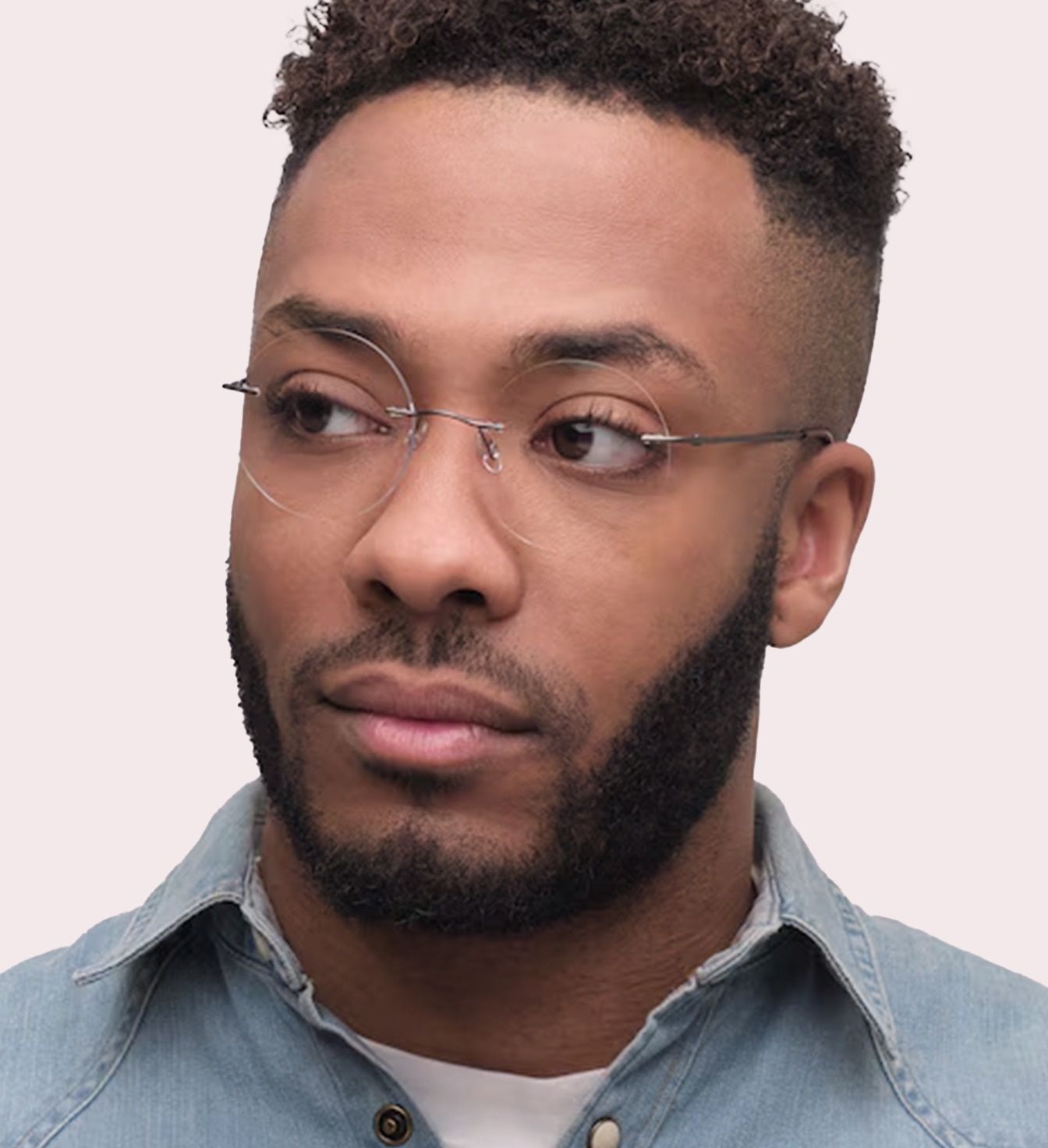 A close-up of a young man with short curly hair, shaved at the sides. He is wearing rimless round glasses and a blue denim shirt.