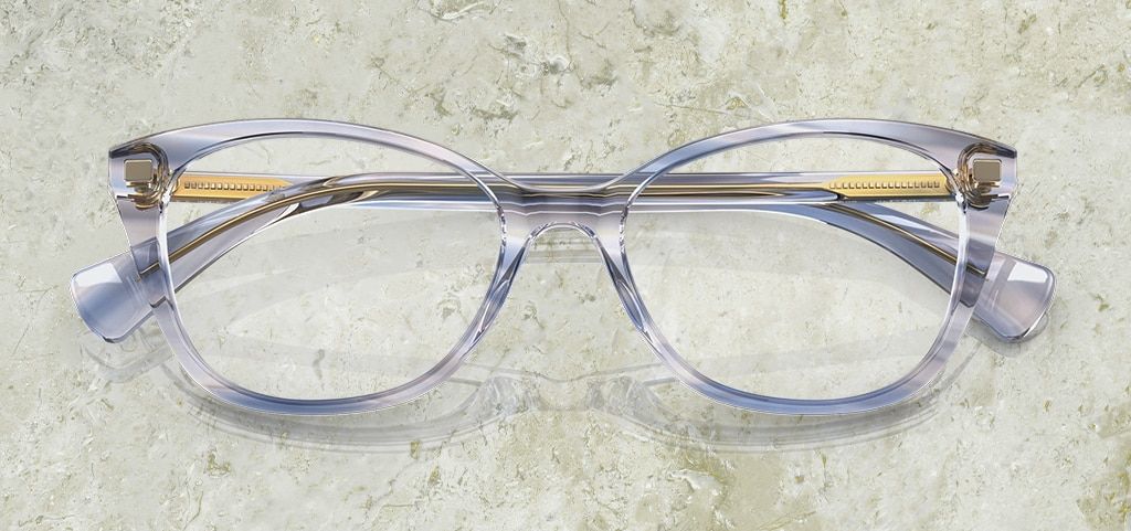 A pair of of acetate pillow-shaped Ralph by Ralph Lauren glasses. They have a transparent blue aesthetic with golden inner temples. The glasses are folded up on a marble background.