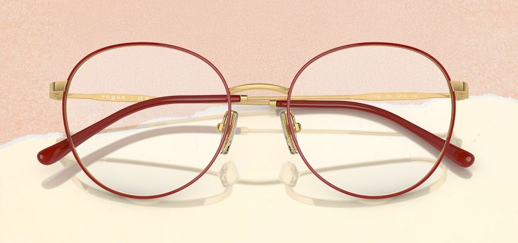 A pair of round, metal Vogue glasses propped open on a paper-torn peach and cream background. The glasses are a gold and red colour with the brand logo adorned along the temples.