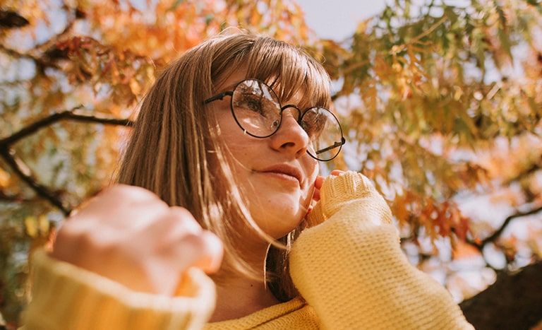 A young woman wearing glasses and a jumper enjoys the autumn sun under a tree