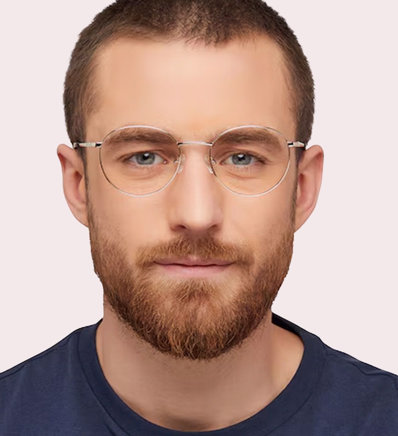 A close-up of a young man with short light-brown hair and a beard. He is wearing round glasses made of silver metal and a blue t-shirt.