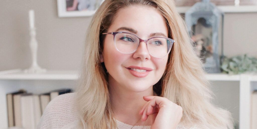 A young, blonde woman wearing cat-eye glasses and smiling while looking off into the distance