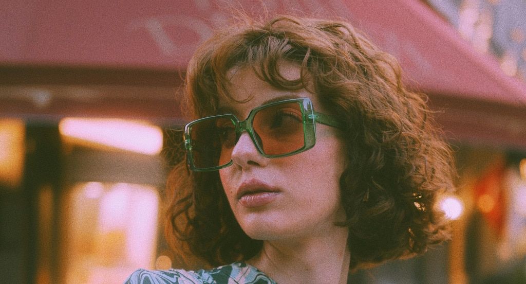 A woman with reddish brown, wavy hair looking sideways. She's wearing large square sunglasses with a green frame and red lenses.