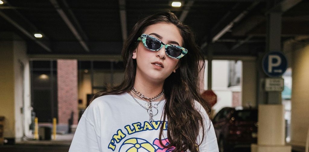 A young woman in a colourful t-shirt and with chains draped around her neck wearing colourful cat-eye sunglasses