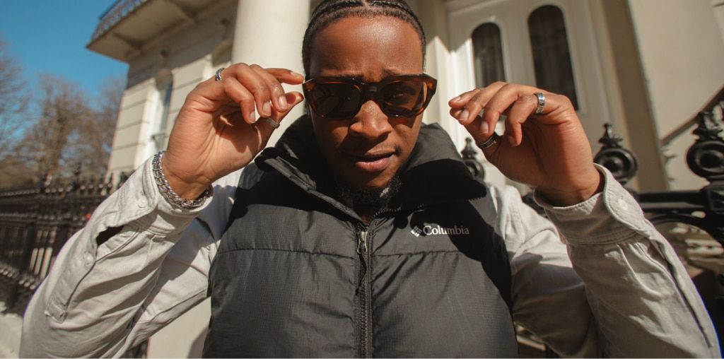 A man with braided hair wearing tortoiseshell sunglasses and a black puffer vest adjusts his frames while standing outdoors in bright sunlight in front of a building with white columns and a black iron fence.