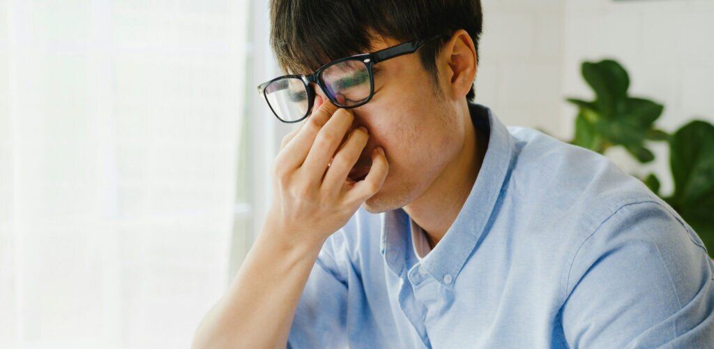 A man with short black hair, wearing a light-blue shirt and rubbing tired, dry eyes under a pair of black square glasses in a bright indoor setting.