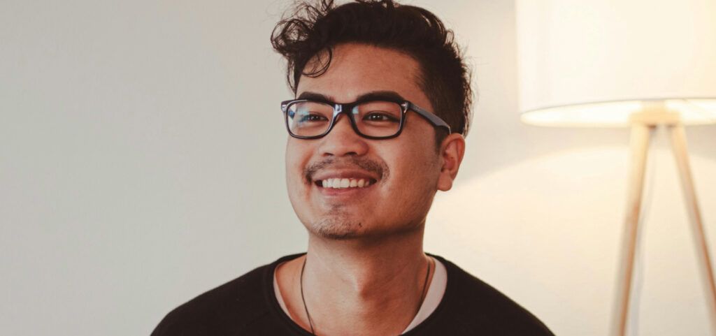 A portrait shot of a young man with short, curly dark-coloured hair. He is sitting and smiling in a bright indoor setting. The man is wearing black rectangular glasses and a dark top. A tall cream-coloured lamp is visible to one side of him.