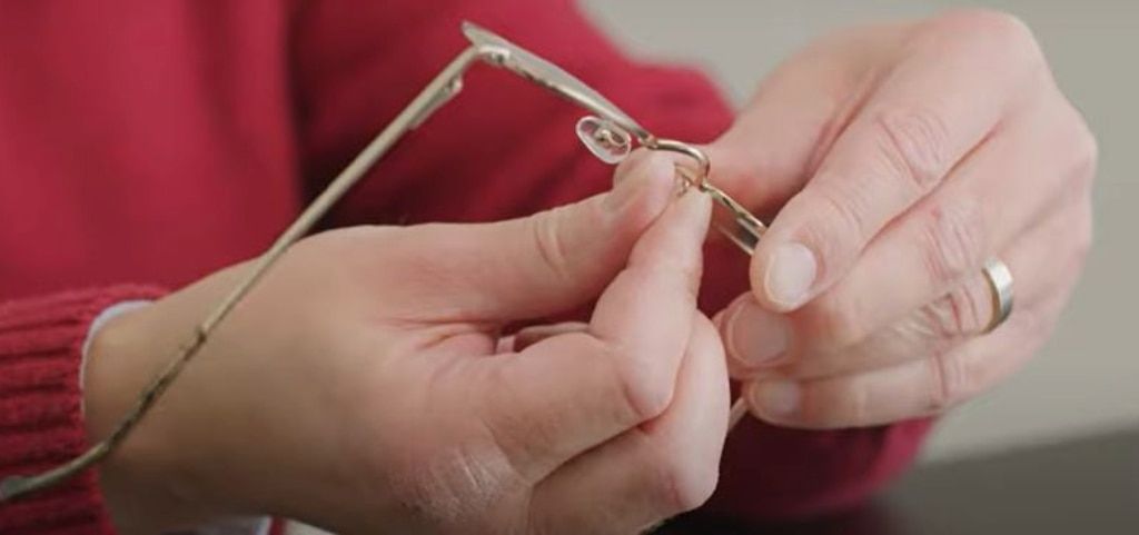 A close up of two hands adjusting the nose pads of a pair of gold metal glasses. The person is wearing a red jumper and has a silver wedding ring.