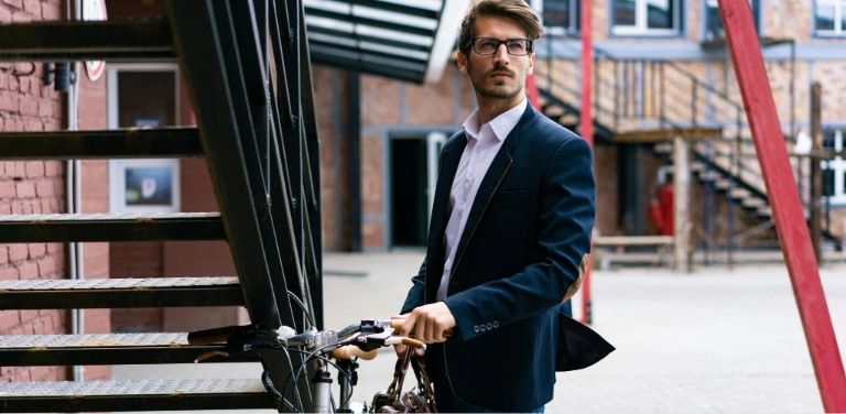 A man with short brown quaffed hair wearing rectangular glasses and a smart navy suit holding a brown briefcase stands beside a bicycle in an urban setting. Looking into the distance, he is positioned near an outdoor staircase and modern buildings, illust