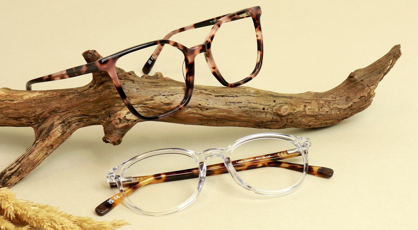 Two pairs of glasses positioned around a wooden branch on a vanilla-coloured background. The frame sitting on the branch has a bold square shape and comes in a pink tortoiseshell colour. The frame positioned in front of the branch is phantos-shaped and cl