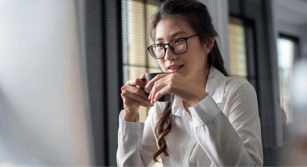 Smiling young woman with long dark hair partly tied back, wearing a white blouse and dark rectangular Asian-fit glasses, holding a grey mug in a bright, modern indoor setting.