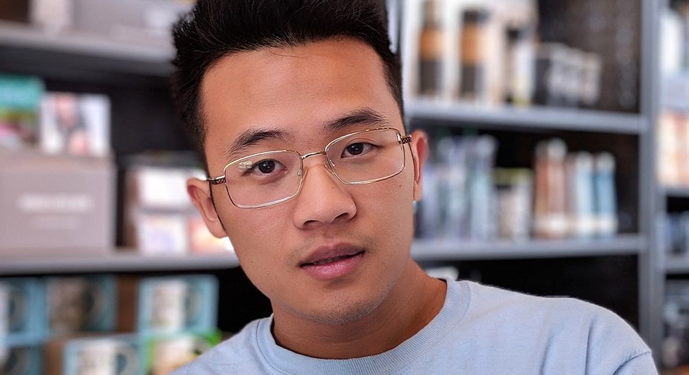 Man with short spiked dark hair wearing a white t-shirt and angular metal Asian-fit glasses, with blurred library bookshelves in the background.