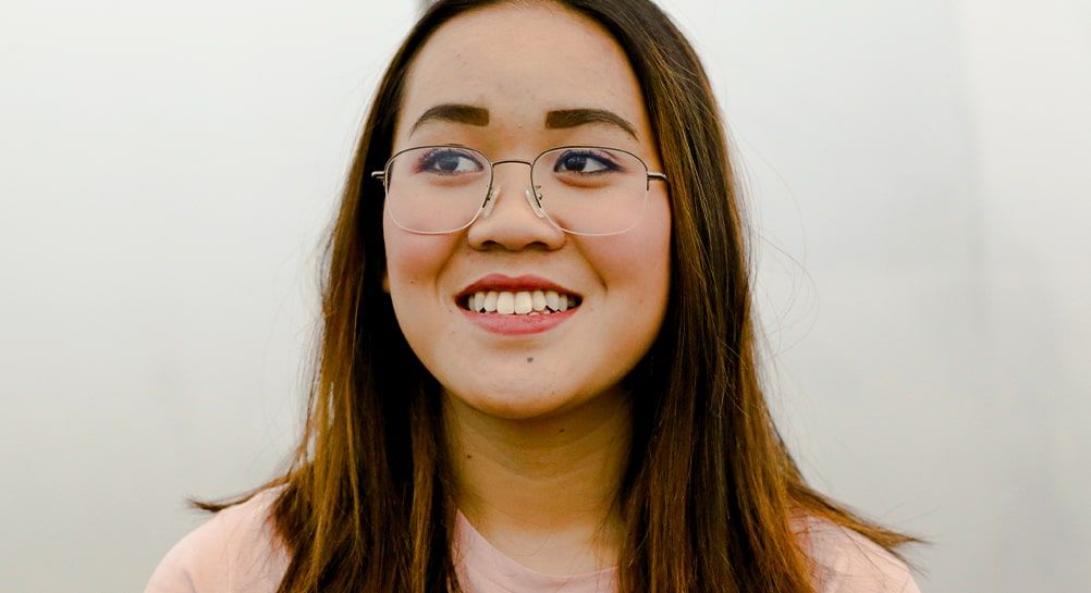 Smiling woman with dark hair and light highlights, wearing a pink top and semi-rimless metal Asian-fit glasses against a plain background.
