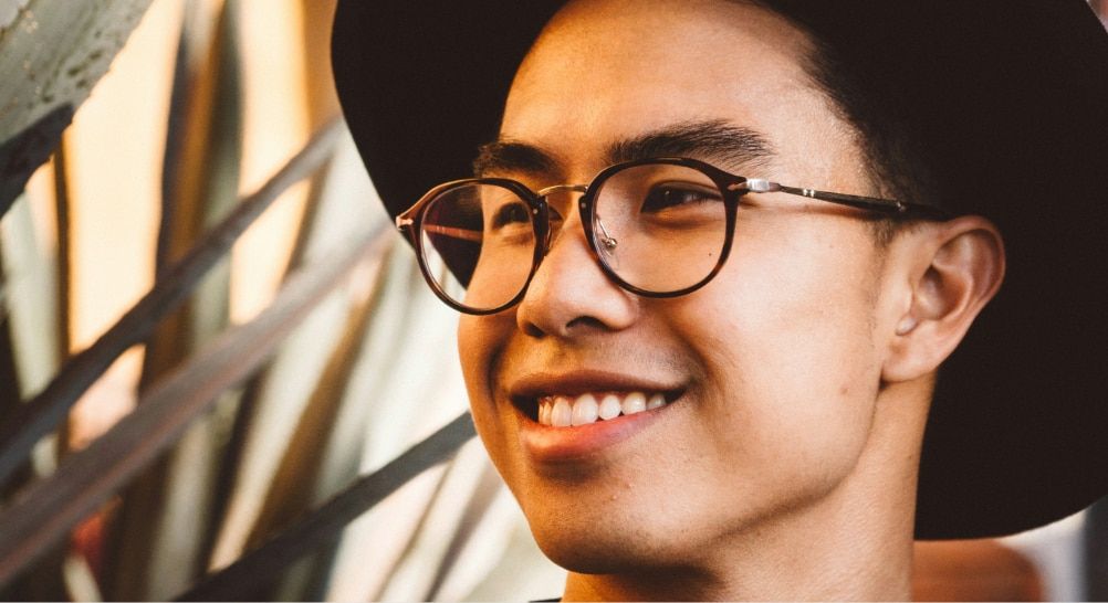 Close-up of a young man with short dark hair wearing dark round Asian-fit glasses with metal accents and a wide-brim hat, looking off to the side outdoors with a neutral expression.