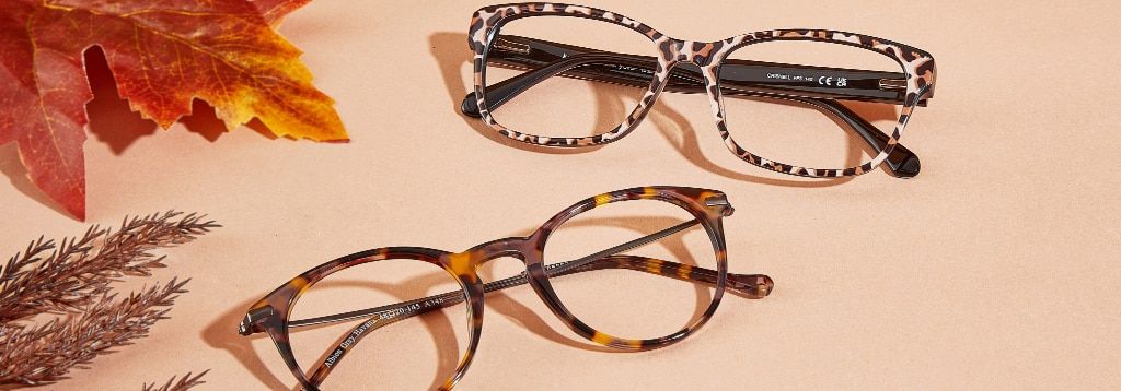 Two frames folded up together with orange leaves and heather peeking from the edges. The acetate frame at the top comes in a bold leopard print while the frame below it is a retro-style round frame with metal temples and a grey Havana front.