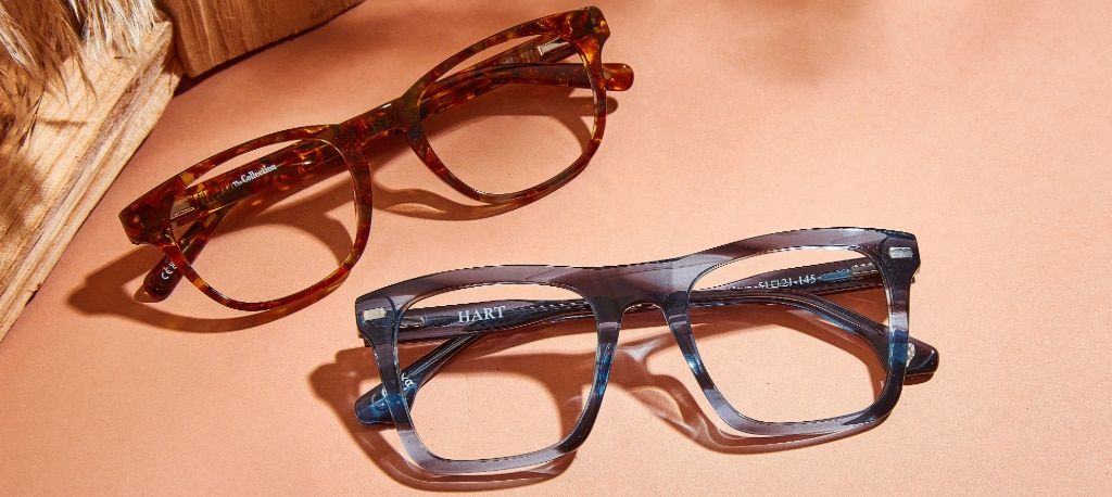 Two acetate frames folded up together with two blocks of wood and barley decorating the corner. One frame is a simple round acetate design in a bright golden Havana and the other is an oversized rectangular frame with a dreamy blue horn aesthetic.