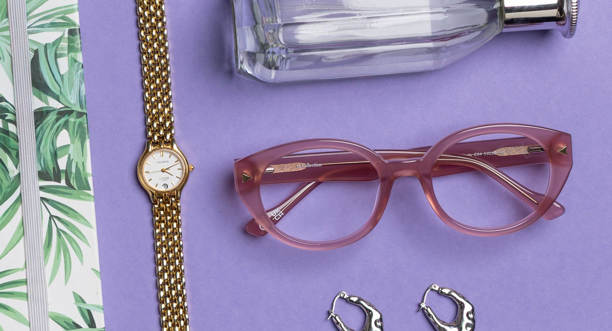 At the centre of a lavender background rests a pair of milky pink oval frames, accompanied by a gold metal wristwatch. Around the edges, glimpses of essentials appear—a white notebook with a green leaf print, gold hoop earrings and a clear perfume bottle.