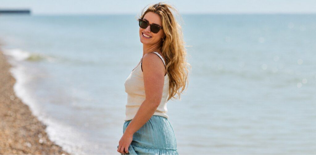 Woman with long wavy blonde hair wearing a cream vest top and light-blue maxi skirt styled with square Seaglass Sands London Retro Balham prescription sunglasses smiling on a sunny beach.